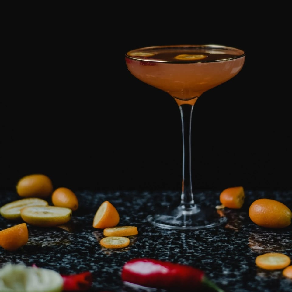 A vibrant orange cocktail in a coupe glass, surrounded by kumquats and red chilies on a dark surface.