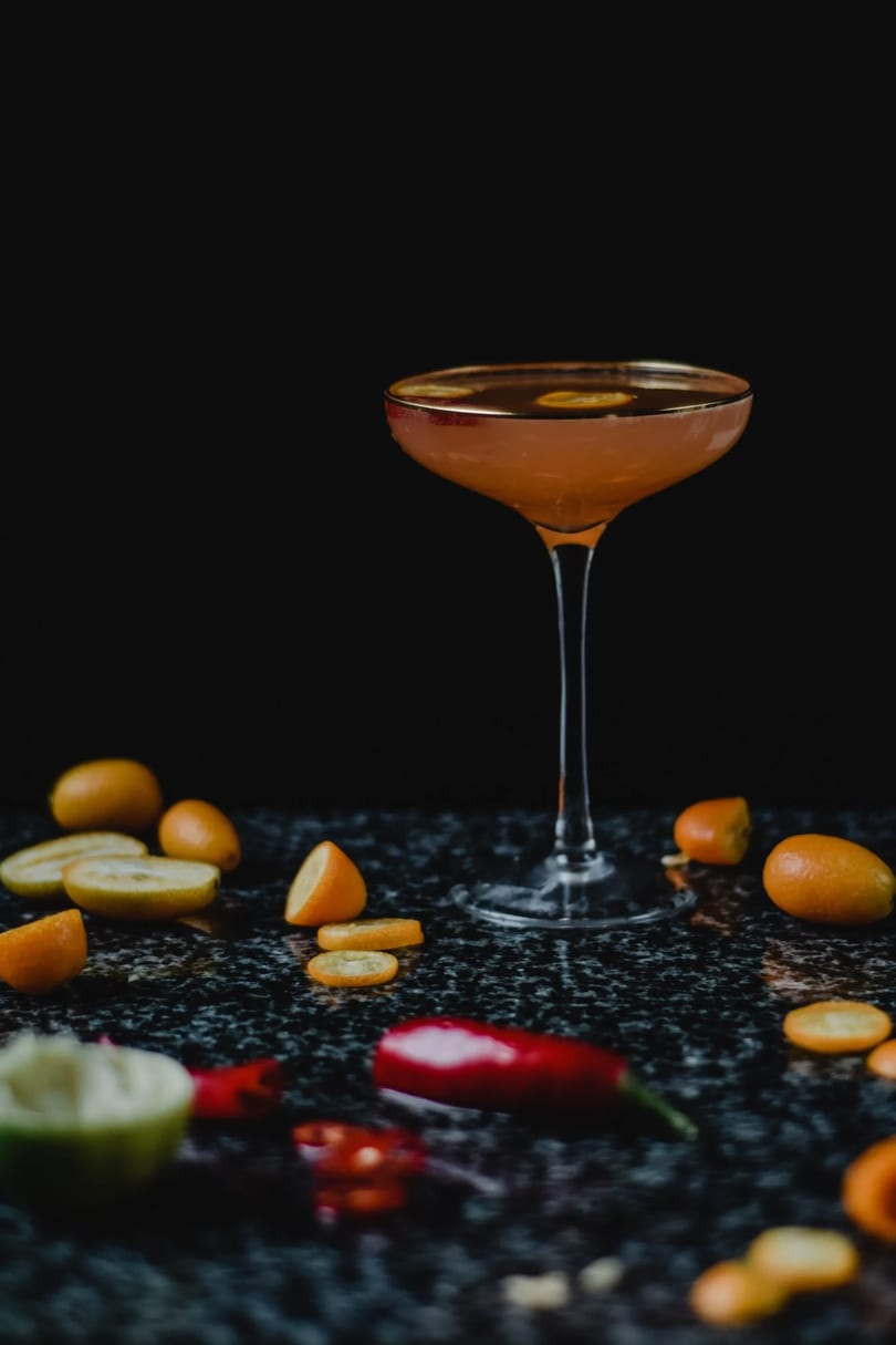 A vibrant orange cocktail in a coupe glass, surrounded by kumquats and red chilies on a dark surface.