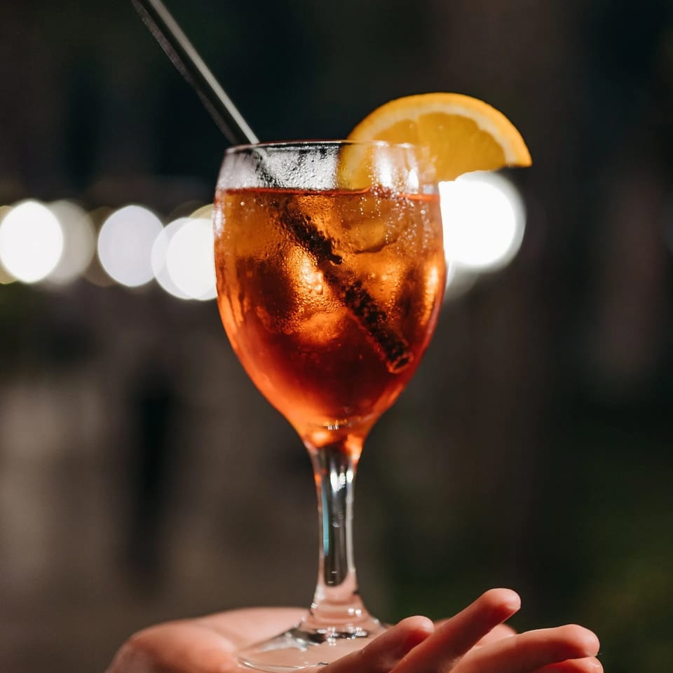 Hand holding a glass of Aperol Spritz with an orange slice and straw, against blurred lights.