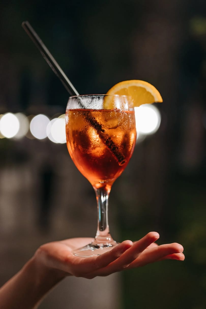 Hand holding a glass of Aperol Spritz with an orange slice and straw, against blurred lights.