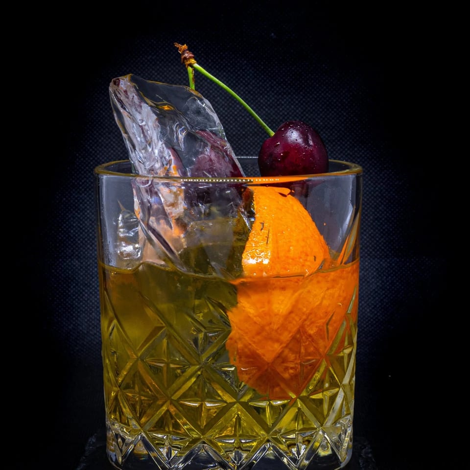 An Old Fashioned cocktail in a cut glass, garnished with an orange peel, ice, and a cherry.