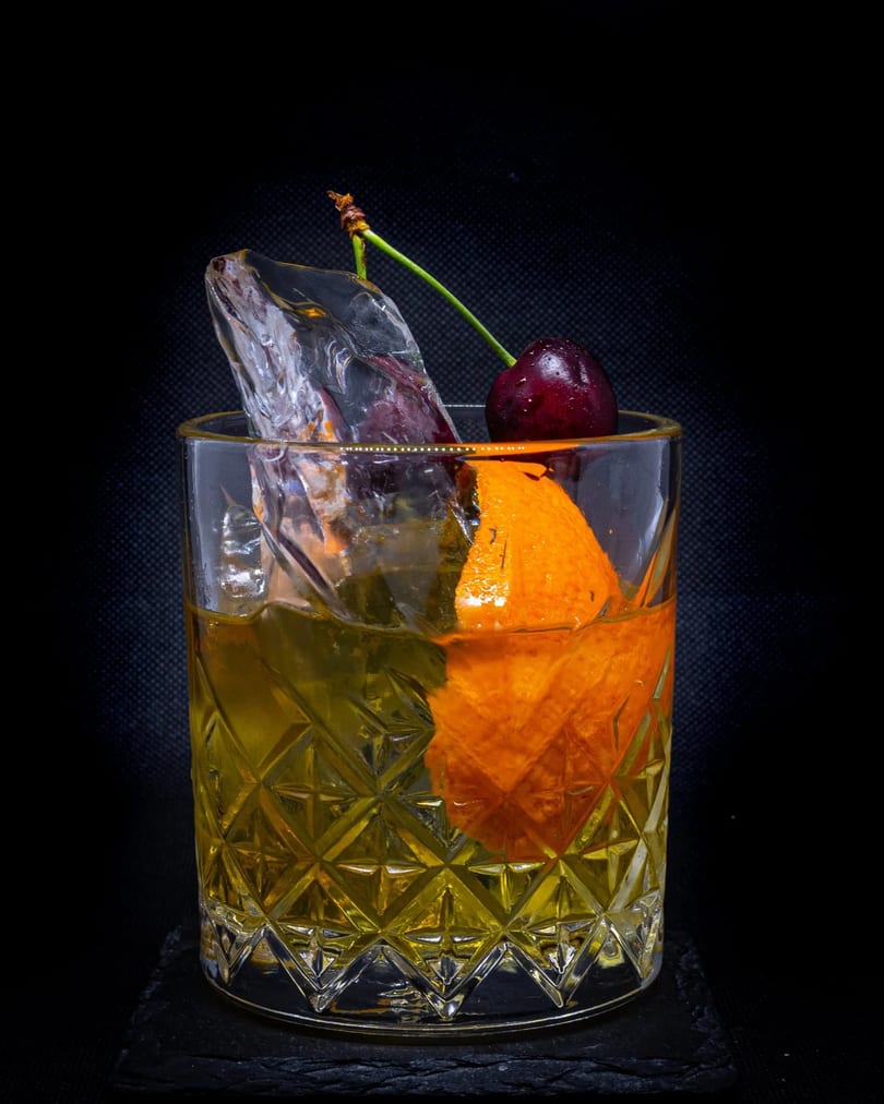An Old Fashioned cocktail in a cut glass, garnished with an orange peel, ice, and a cherry.